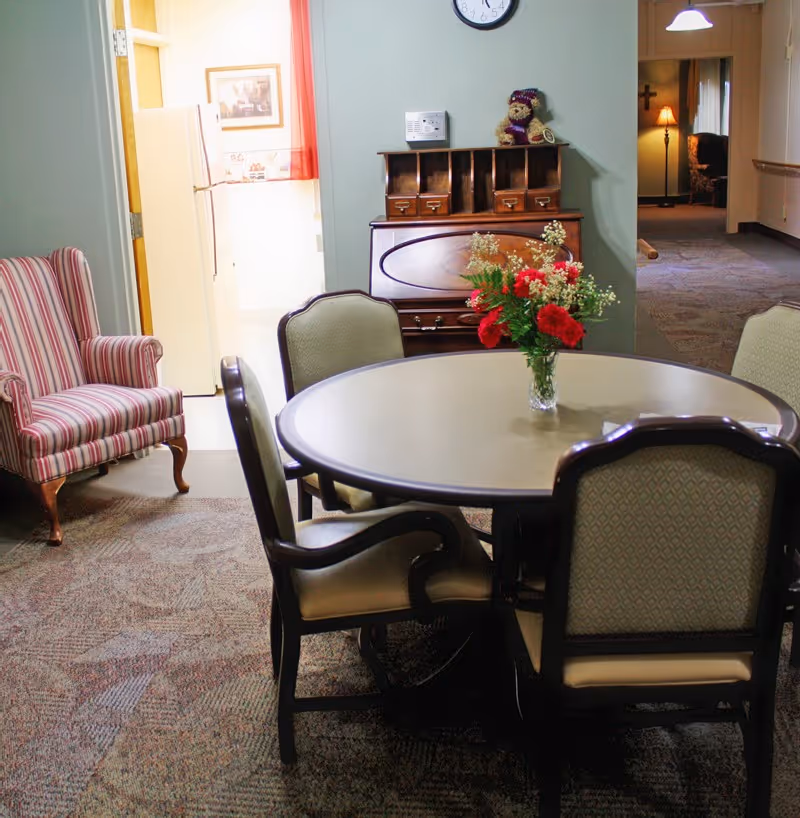 A cozy interior room with a round table surrounded by four upholstered chairs. A vase with red and white flowers is placed on the table. To the left, there is a striped armchair. In the background, a wooden cabinet with cubbyholes and a teddy bear on top is against a green wall with a clock above it. A doorway leads to a kitchen area with a refrigerator, and a hallway extends to another room with a lamp and a cross on the wall.