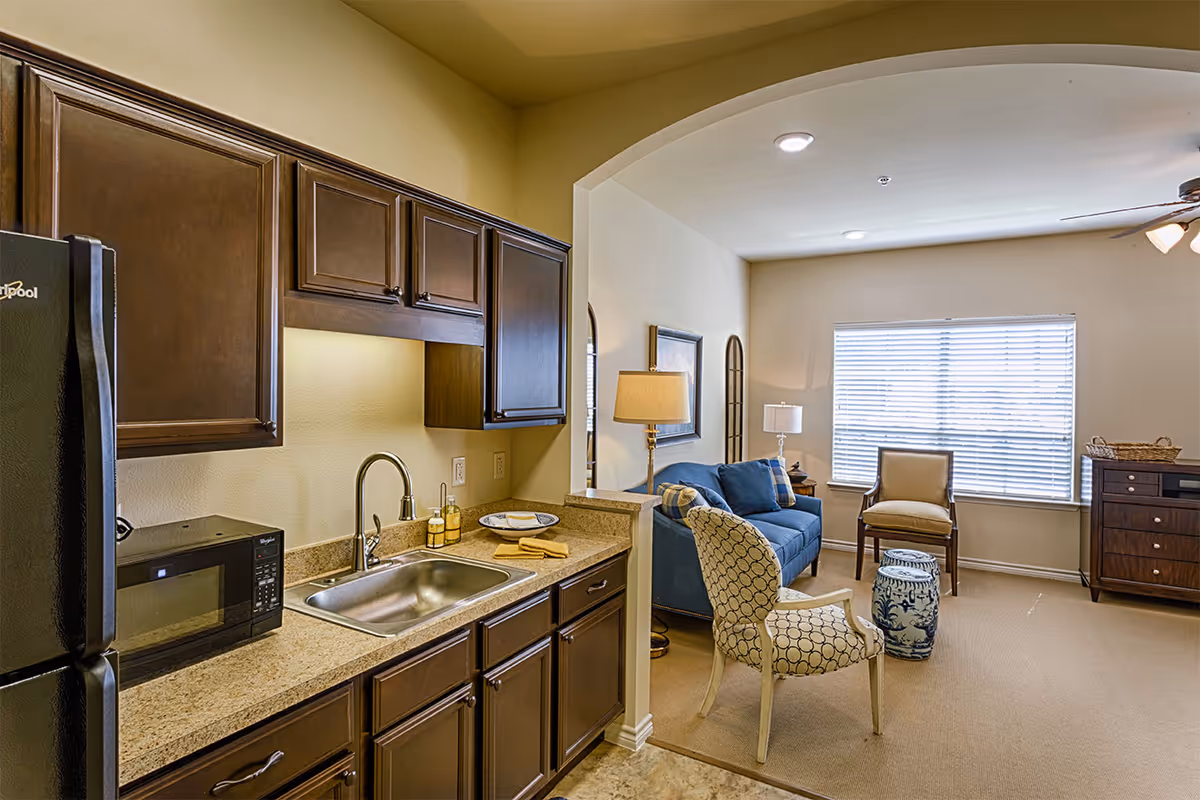 Open-plan apartment showing a small kitchen with dark wood cabinets and sink opening to a living area with a blue sofa, chairs, and a large window.
