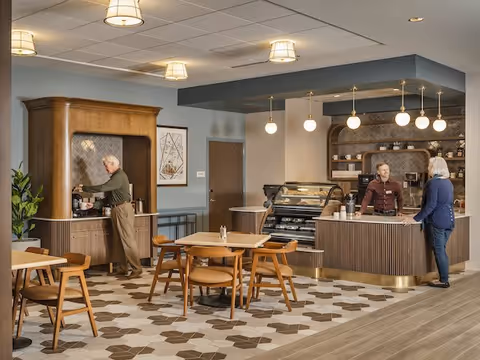 A modern cafe area inside a senior living facility with wooden tables and chairs. An elderly man is using a coffee machine on the left, while two women are conversing at the counter on the right. The space features pendant lighting, a display case, and decorative shelving behind the counter.