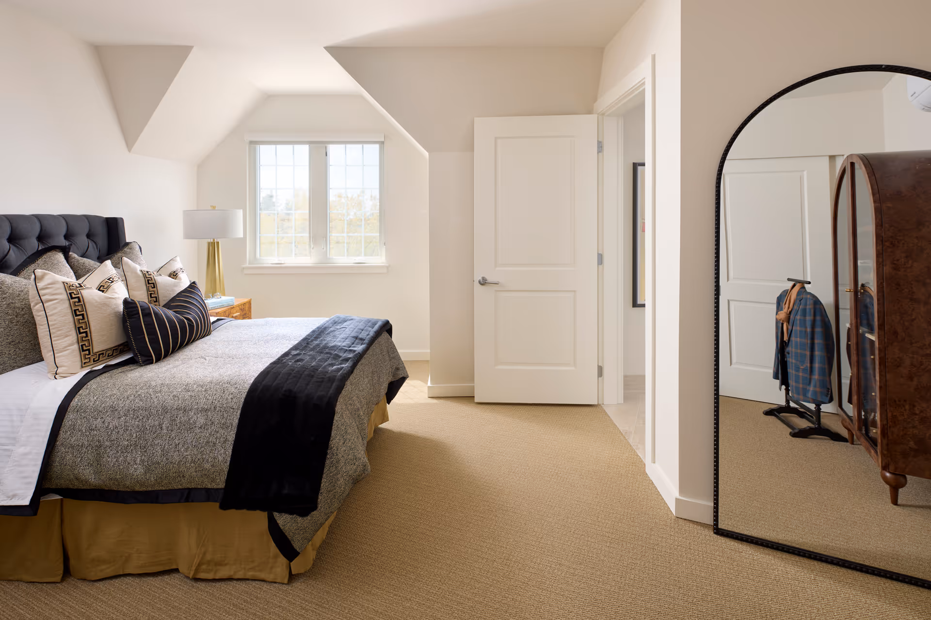 A bright bedroom with a large bed featuring multiple decorative pillows and a black throw blanket. There is a window with white blinds letting in natural light, a bedside table with a gold lamp, and a large arched mirror reflecting part of the room including a wooden cabinet and a clothing rack with a jacket and scarf. The walls are painted white and the carpet is beige.