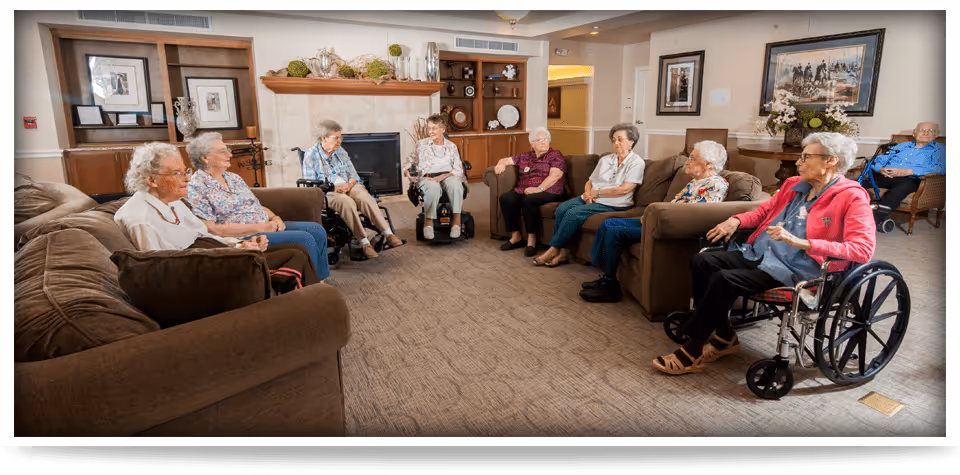 A group of elderly individuals sitting in a circle in a cozy living room area with brown sofas and armchairs. The room features a fireplace, wooden shelves with decorative items, framed artwork on the walls, and carpeted flooring. Some individuals are seated in wheelchairs while others sit on chairs and sofas, engaging in a social activity.