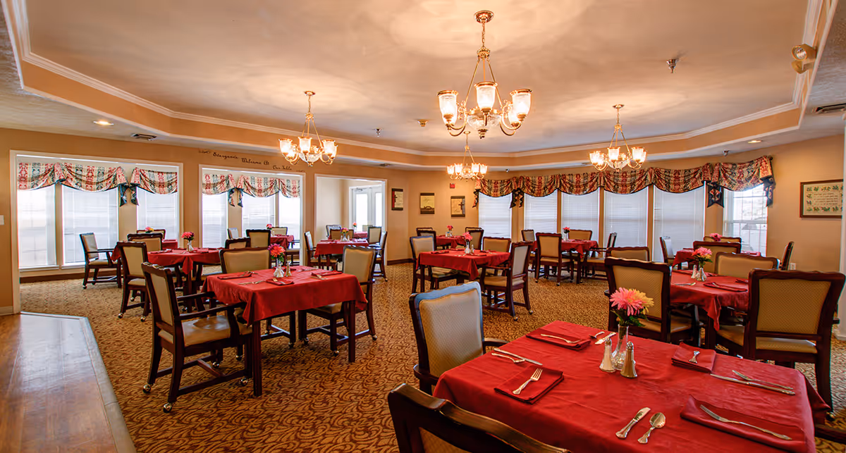 A dining room with multiple tables covered in red tablecloths, each set with silverware, napkins, and small flower vases. The room has patterned carpet, beige walls, large windows with floral valances, and several chandeliers hanging from the ceiling providing warm lighting.