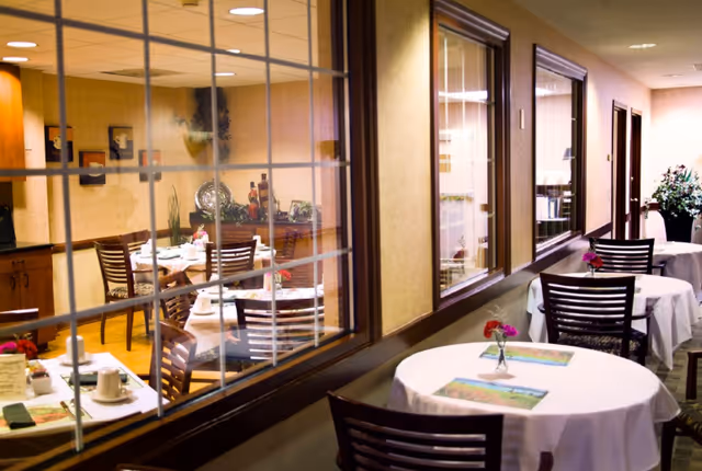 Interior dining area with round tables set with white tablecloths and chairs along a windowed corridor.