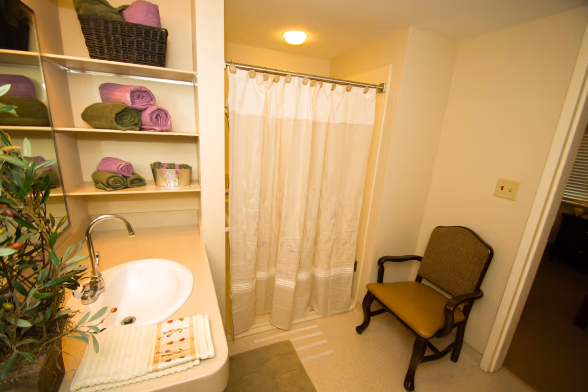 Bathroom with a sink and faucet on the left, shelves holding rolled green and purple towels, a basket, and a small decorative container. A shower with a beige curtain is in the center background. A cushioned chair with wooden arms and legs is positioned against the right wall. The walls are light-colored and the floor is light-toned.