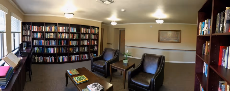 A cozy reading room in a senior living facility with large bookshelves filled with books along the walls, two dark leather armchairs, a coffee table with books on it, and a small side table with a potted plant. The room has beige walls, carpeted floor, and ceiling lights, with windows on the left side letting in natural light.