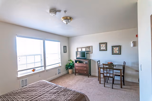 A bright senior living facility room with a bed in the foreground, two large windows with blinds letting in natural light, a small wooden dining table with four chairs, a wooden dresser with a small TV on top, a potted plant in the corner, and three framed pictures on the wall.