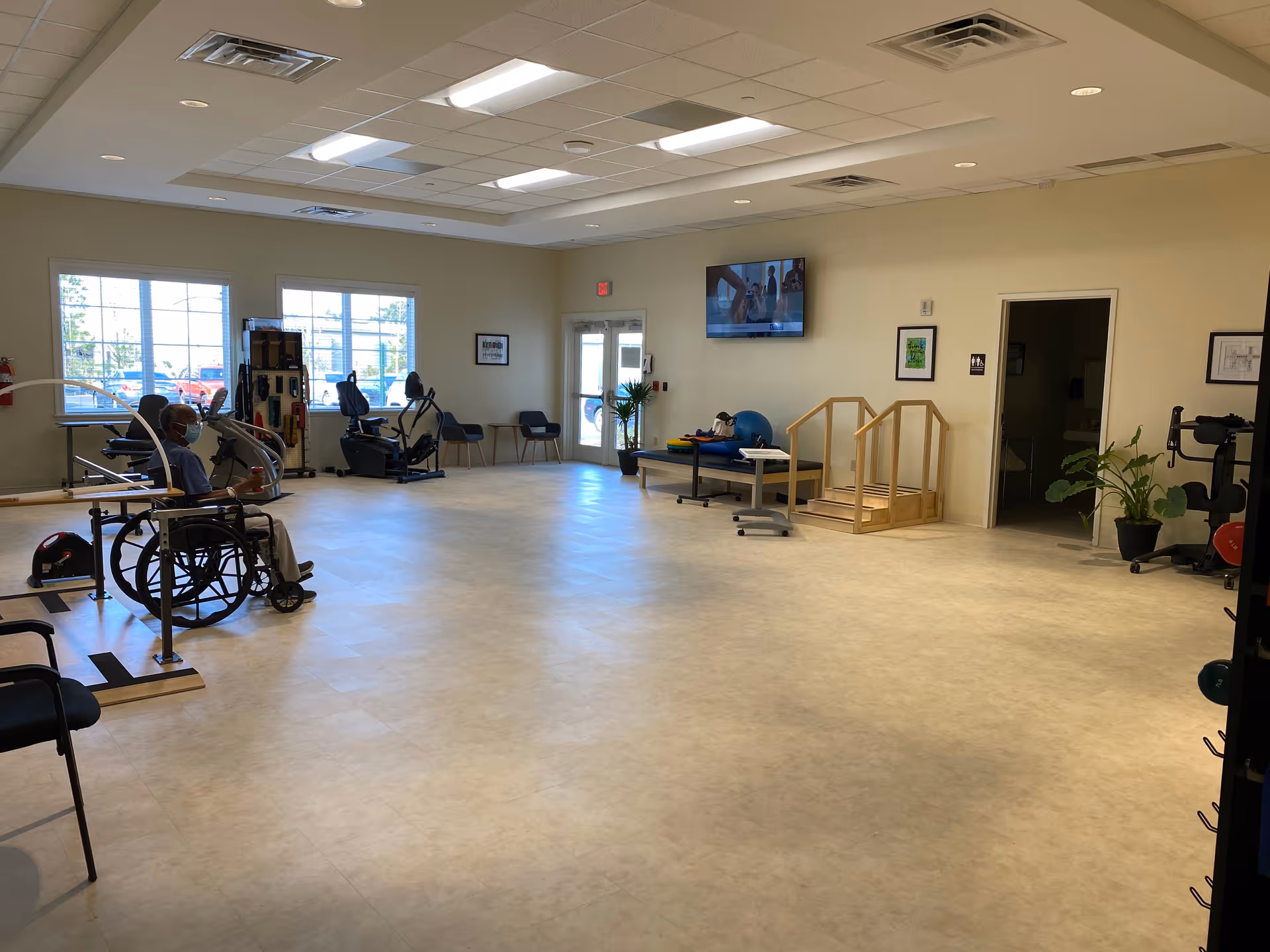 A spacious physical therapy or rehabilitation room in a nursing home with exercise equipment, parallel bars, chairs, and a person in a wheelchair wearing a mask. The room has large windows, a TV mounted on the wall, and a small set of wooden stairs for therapy exercises.