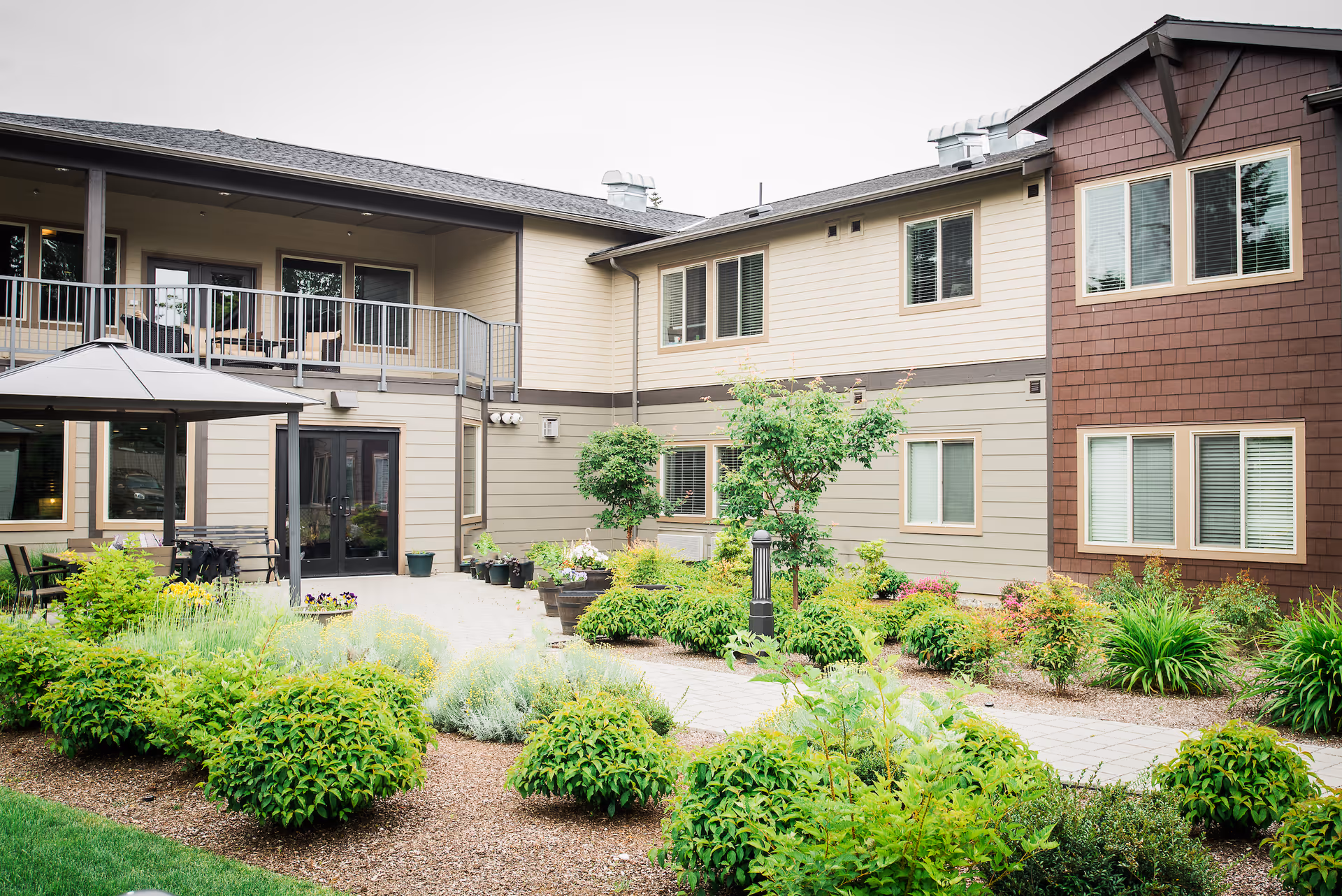 Outdoor courtyard area of a senior living facility with well-maintained garden beds, shrubs, and small trees. The building surrounding the courtyard has two stories with multiple windows and a balcony with outdoor seating. There is a covered gazebo structure and a paved walkway through the garden.