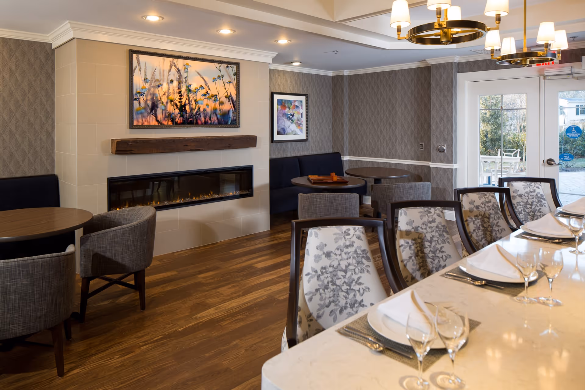 A cozy dining and lounge area featuring a modern electric fireplace with a wooden mantel and a framed floral artwork above it. The room has wooden flooring, patterned wallpaper, and recessed ceiling lights. There are round tables with upholstered chairs and a long dining table set with plates, napkins, and glassware. A glass door leads to an outdoor patio area.