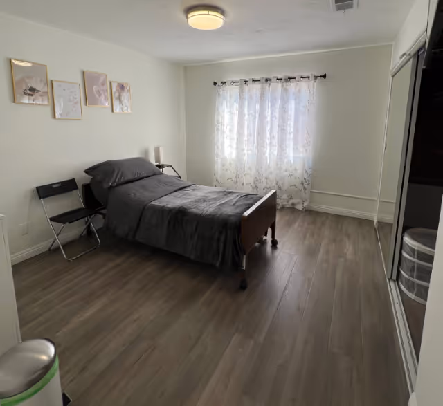 A simple bedroom with a single bed covered in dark gray bedding, a black folding chair beside the bed, three framed pictures on the wall, a window with sheer floral curtains letting in natural light, and a mirrored closet door on the right side.
