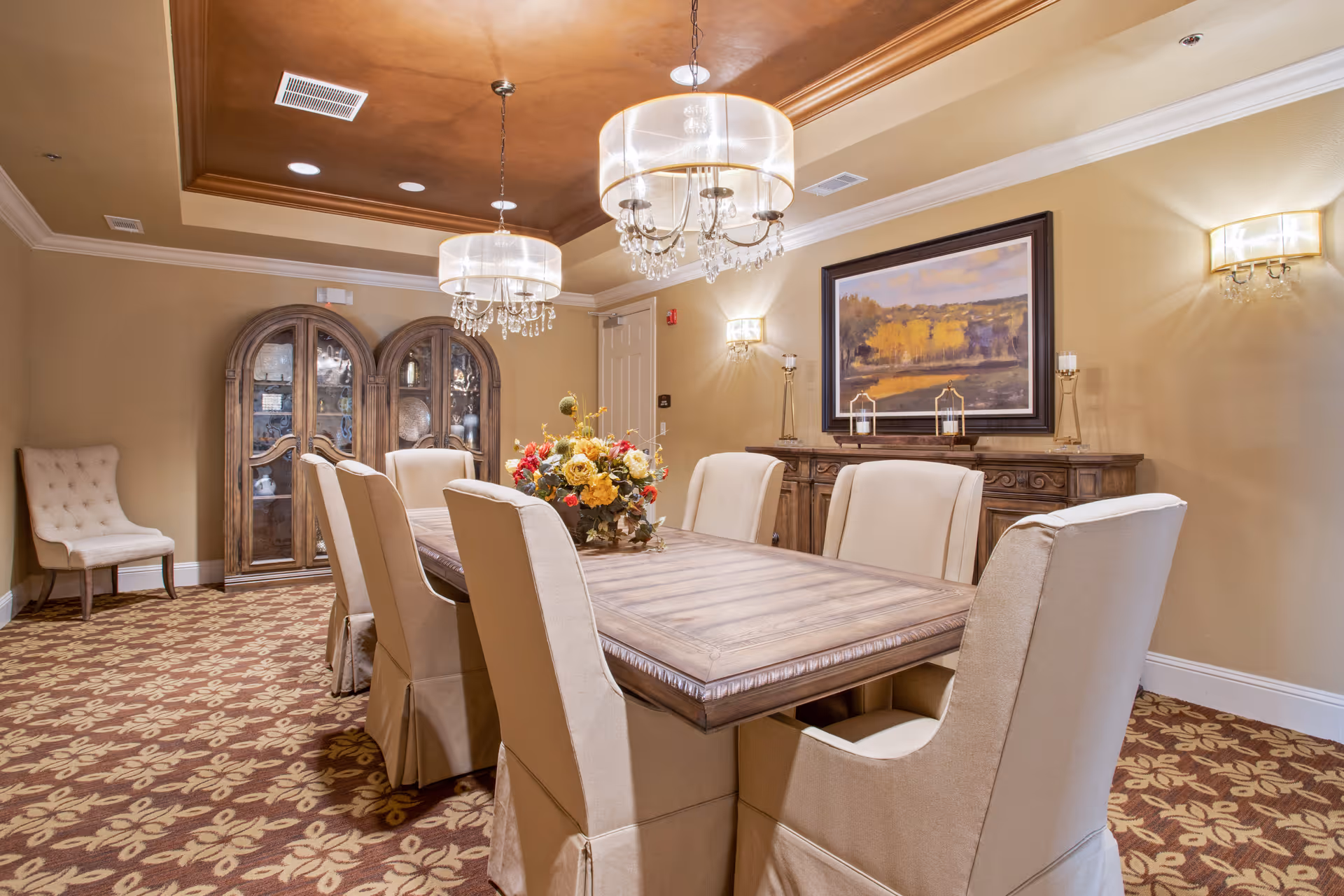 A well-lit dining room with a long wooden table surrounded by eight beige upholstered chairs. The table has a floral centerpiece. The room features a patterned carpet, two chandeliers with crystal accents hanging from a recessed ceiling, a wooden sideboard with decorative items, and a large framed landscape painting on the wall. There is also a tufted beige chair in the corner and a wooden cabinet with glass doors.