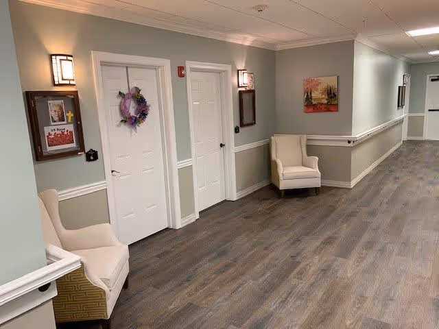 A clean and well-lit hallway in a senior living facility with light green walls, white doors, and wood-look flooring. Two beige armchairs are placed along the walls, and there are framed pictures and a wreath on the doors. Wall-mounted lights provide soft illumination.