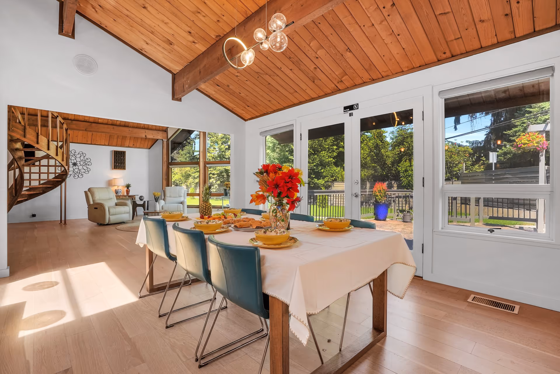 A bright dining room with a wooden ceiling and light wood flooring. A rectangular dining table covered with a white tablecloth is set with yellow bowls, plates, and a vase of red and orange flowers. Blue chairs surround the table. Large windows and glass doors provide a view of a green outdoor garden area. In the background, there is a living area with two beige recliner chairs and a wooden spiral staircase.