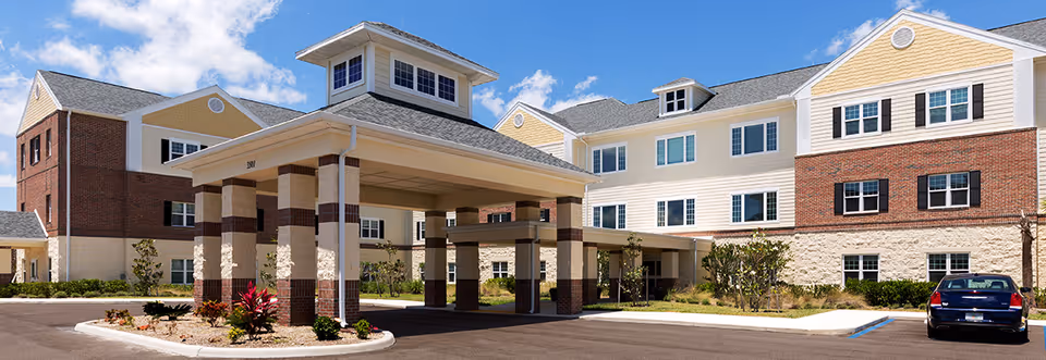 Front exterior of a multi-story senior living facility with a covered porte-cochère entrance, landscaped beds, and a parked car.