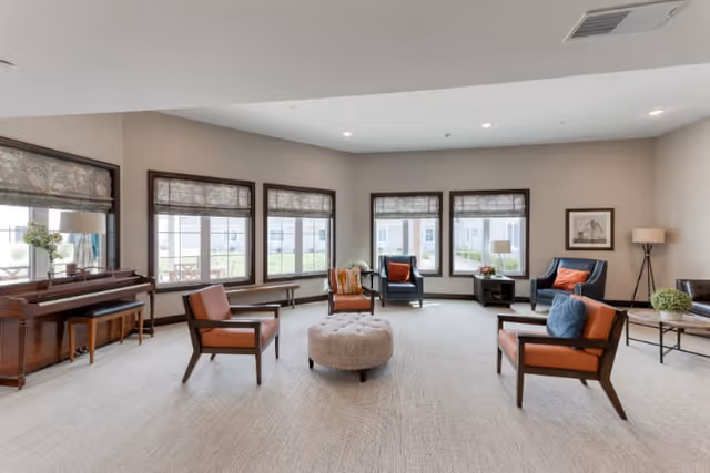 A spacious senior living common area with large windows allowing natural light to fill the room. The room features several modern armchairs with orange cushions arranged around a round tufted ottoman in the center. There is a piano on the left side with a bench, a floor lamp in the corner, and a coffee table with a plant on the right. The walls are painted light beige, and the carpet is light-colored.