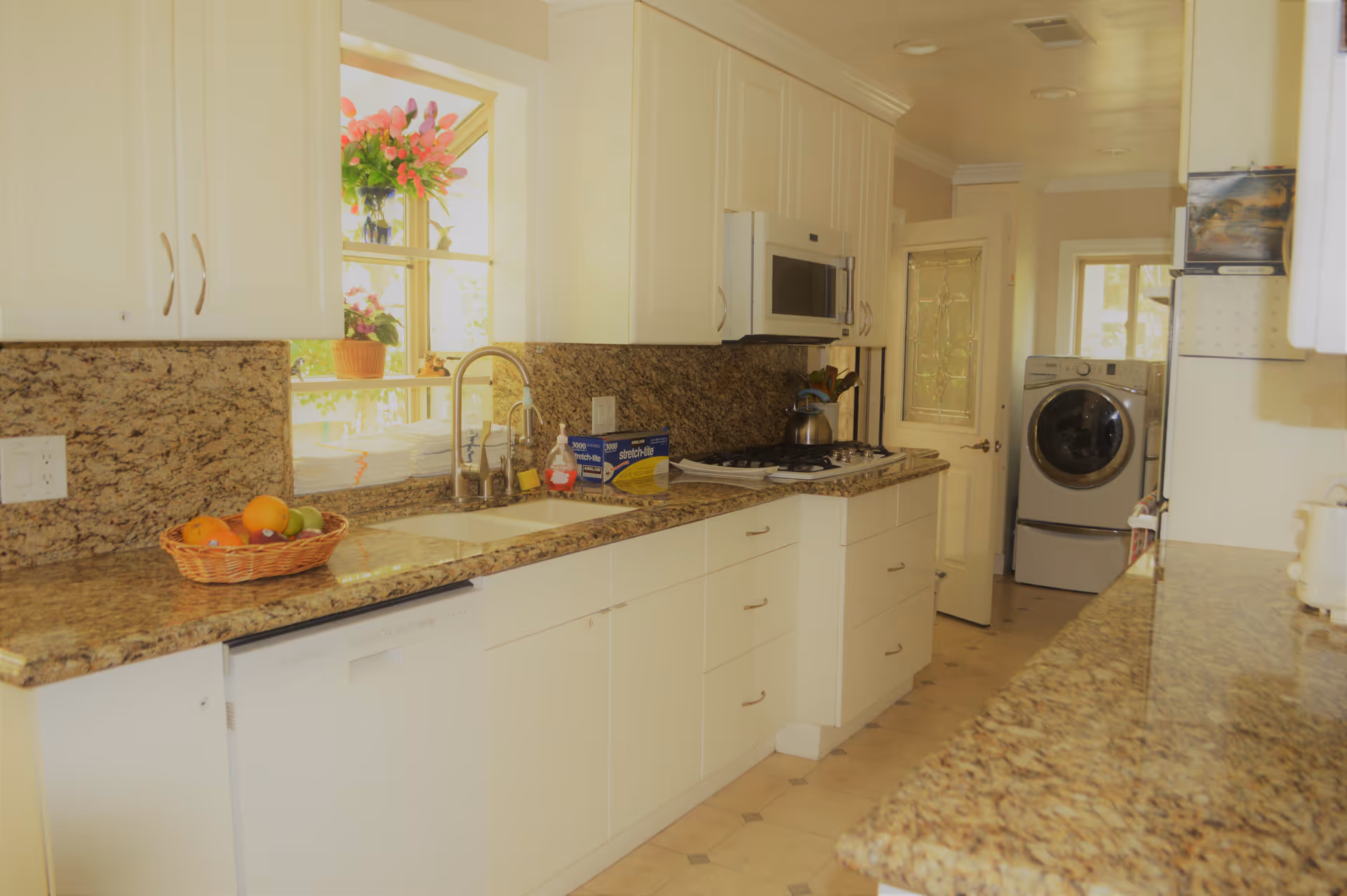 A bright kitchen with white cabinets and granite countertops. There is a sink under a window with flowers on the windowsill, a basket of fruit on the counter, a microwave above a stove, and a washing machine visible in the background near a door with decorative glass.