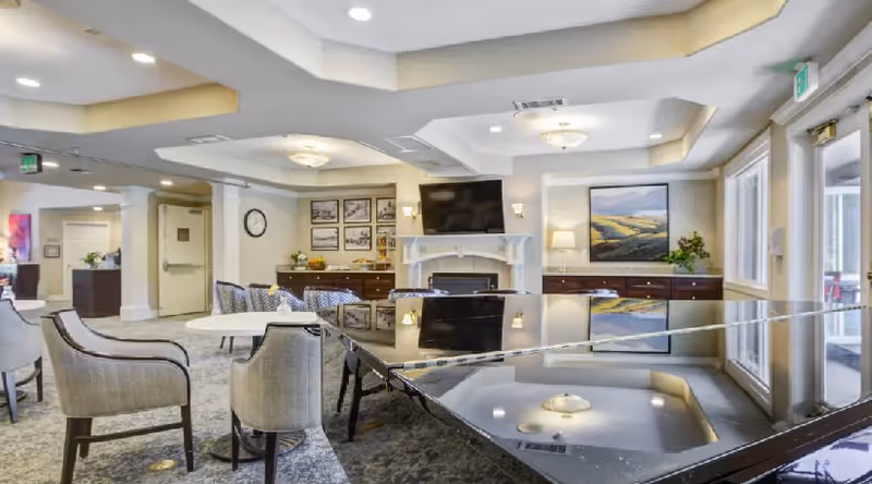 A bright and spacious common area in a senior living facility featuring a glossy black grand piano in the foreground, several upholstered chairs and tables, a wall-mounted flat-screen TV above a white fireplace, framed artwork on the walls, and large windows allowing natural light to fill the room.