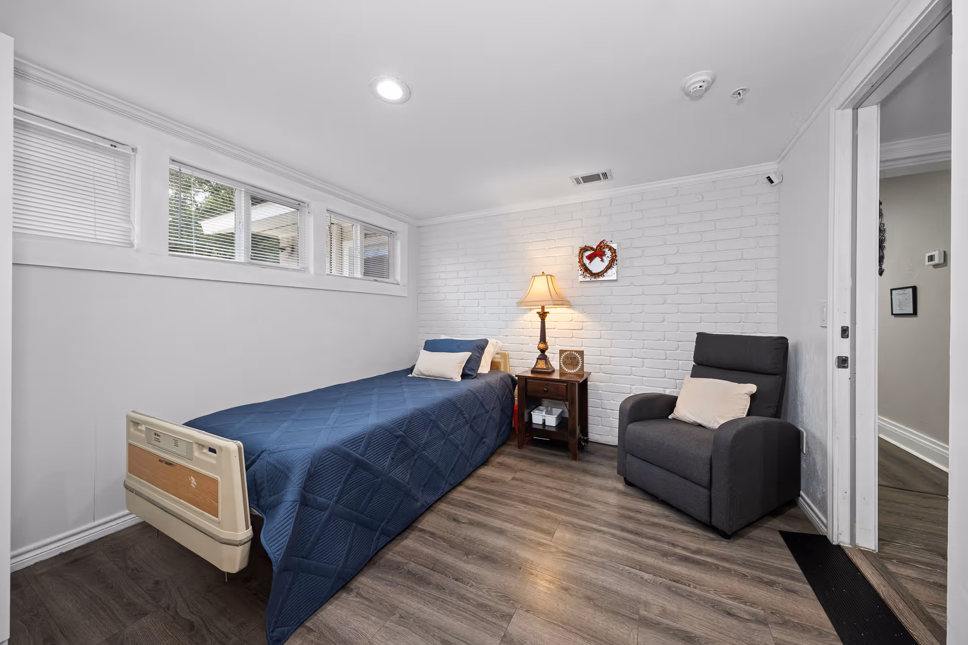 A small, clean bedroom with a single bed covered in a blue quilt, a wooden nightstand with a lamp and decorative items, a gray armchair with a pillow, and white brick walls. There are three small windows near the ceiling letting in natural light, and the floor is wood laminate.