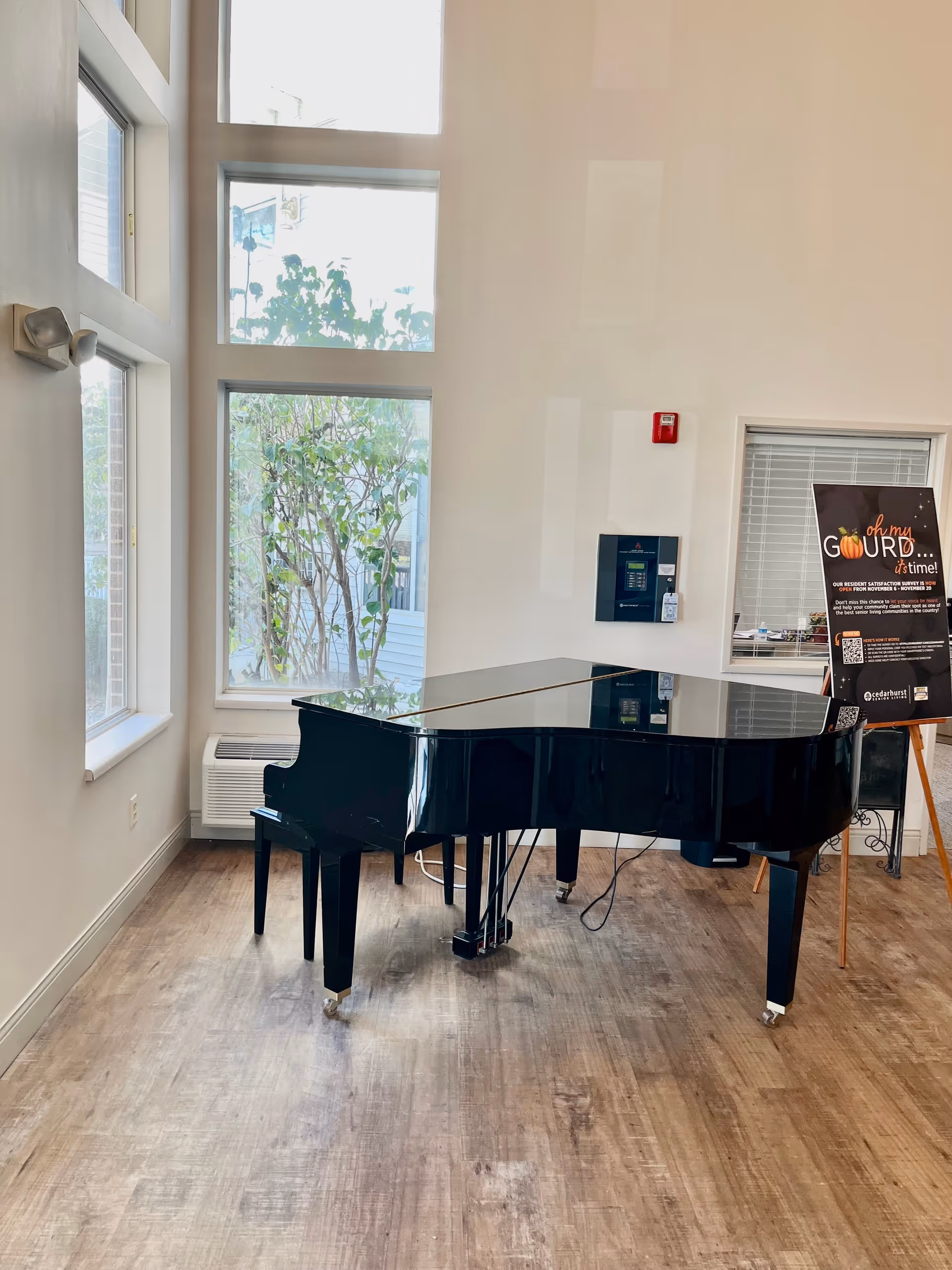 A black grand piano placed on a wooden floor in a bright room with large windows showing greenery outside. There is a wall-mounted device and a fire alarm on the wall behind the piano. To the right, there is a stand with a sign featuring autumn-themed graphics and text.