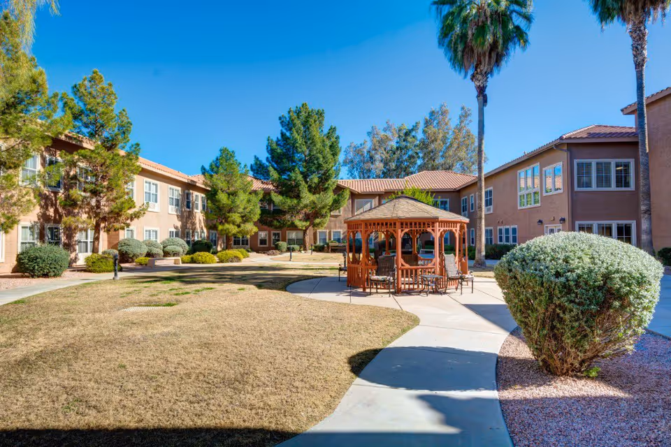 Outdoor courtyard area of Pegasus Landing of Mesa featuring a wooden gazebo with chairs and tables, surrounded by a paved walkway, bushes, trees, and a two-story building with multiple windows under a clear blue sky.