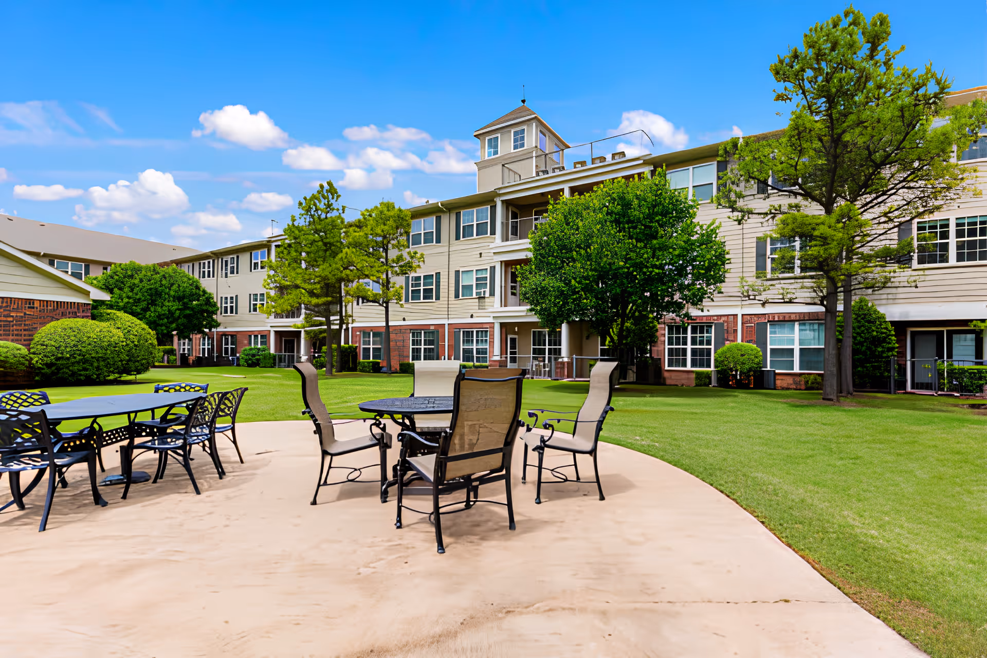Patio area with metal tables and chairs on a paved patio in front of a three-story residential building and grassy courtyard.