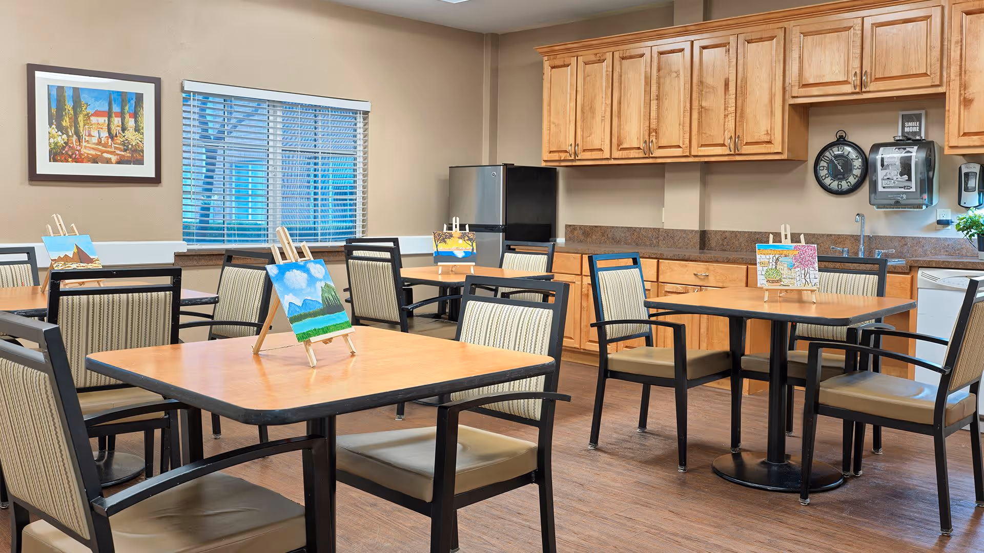 A well-lit activity room with several tables and chairs arranged neatly. Each table has a small easel displaying a colorful painting. The room features wooden cabinets, a refrigerator, a wall clock, a paper towel dispenser, and a window with blinds. A framed painting hangs on the wall.