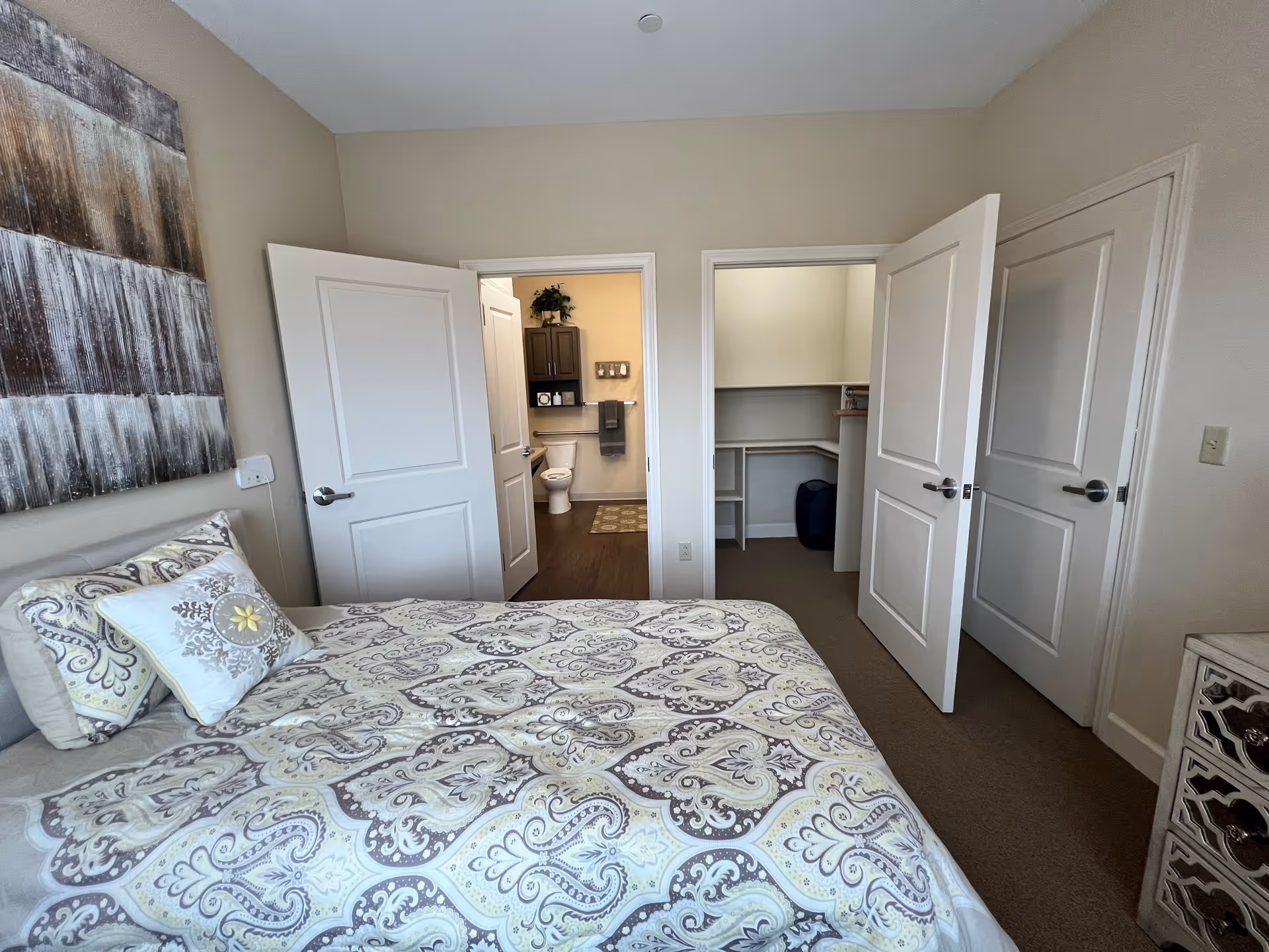 A bedroom with a bed covered in a patterned bedspread and pillows. The room has beige walls and a large piece of abstract art hanging above the bed. There are three white doors visible: one leading to a bathroom with a toilet and cabinet, one leading to a walk-in closet with shelves, and one closed door. A small nightstand with a decorative front is on the right side of the image.