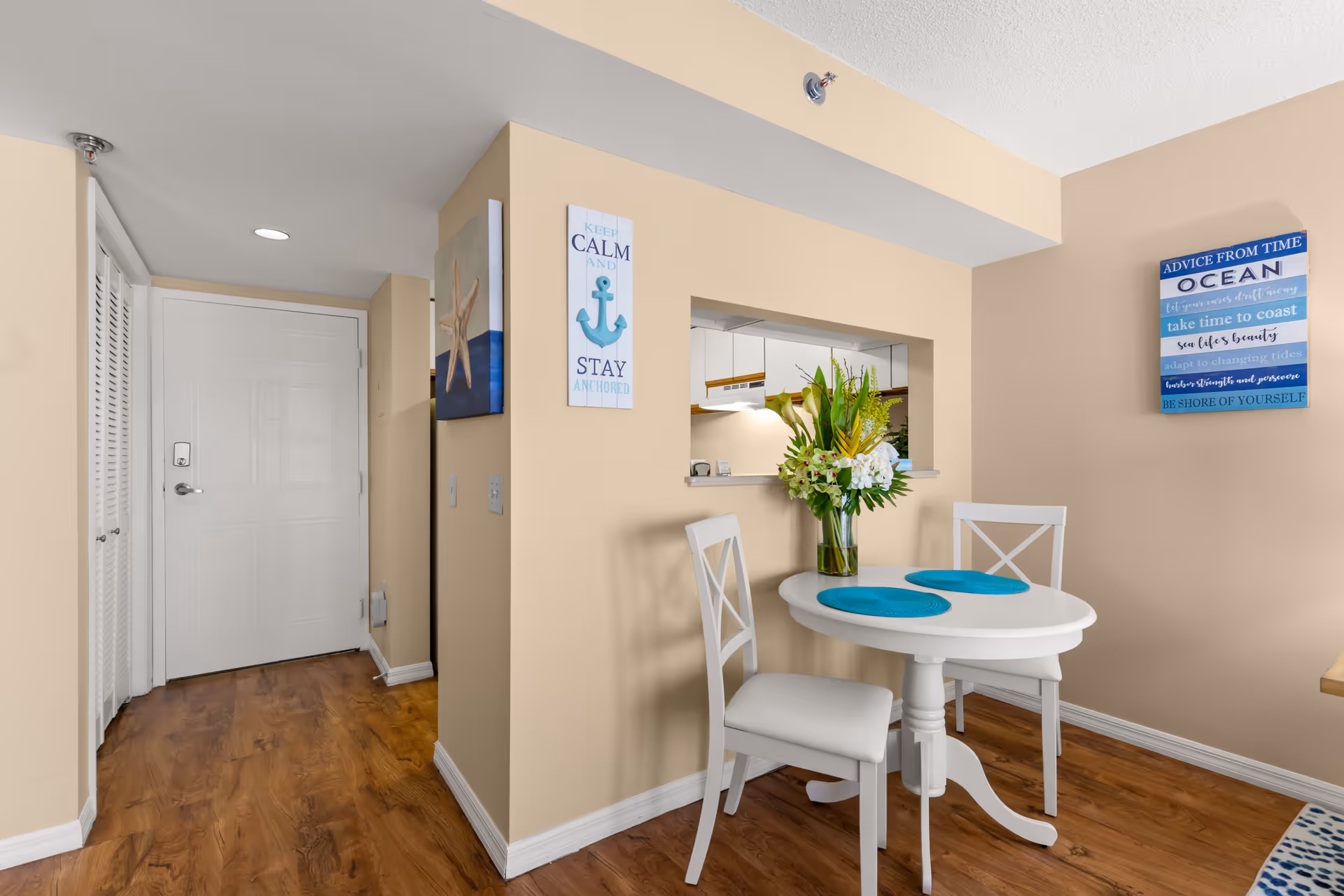 Small dining area with a white round table and two white chairs with blue placemats. A vase with flowers is on the table. The walls are beige with two decorative wall signs, one with an anchor and the other with ocean-themed advice. The floor is wooden, and there is a white door and a kitchen pass-through window in the background.