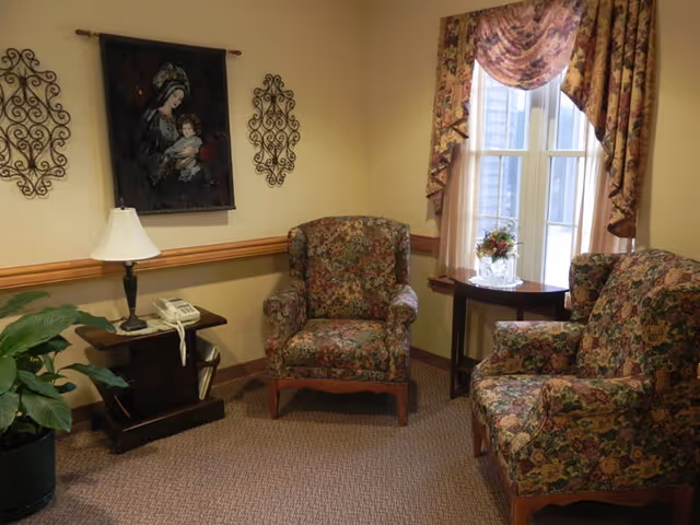 A cozy sitting area with two floral upholstered armchairs, a small wooden side table with a lamp and a telephone, a round table with a floral arrangement near a window with floral curtains, and wall decorations including a tapestry and two metal wall hangings.