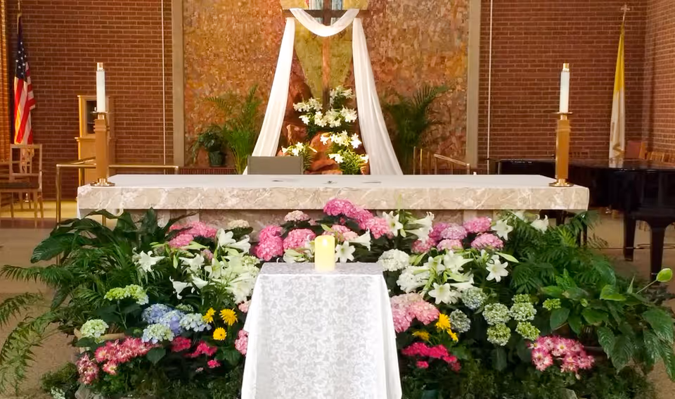 Interior view of a chapel with a stone altar adorned with white cloth and surrounded by colorful flowers including pink, white, and purple blooms. Behind the altar is a wooden cross draped with white fabric, flanked by green plants. American and Vatican flags are visible on either side, along with two tall candles on the altar and a piano to the right.