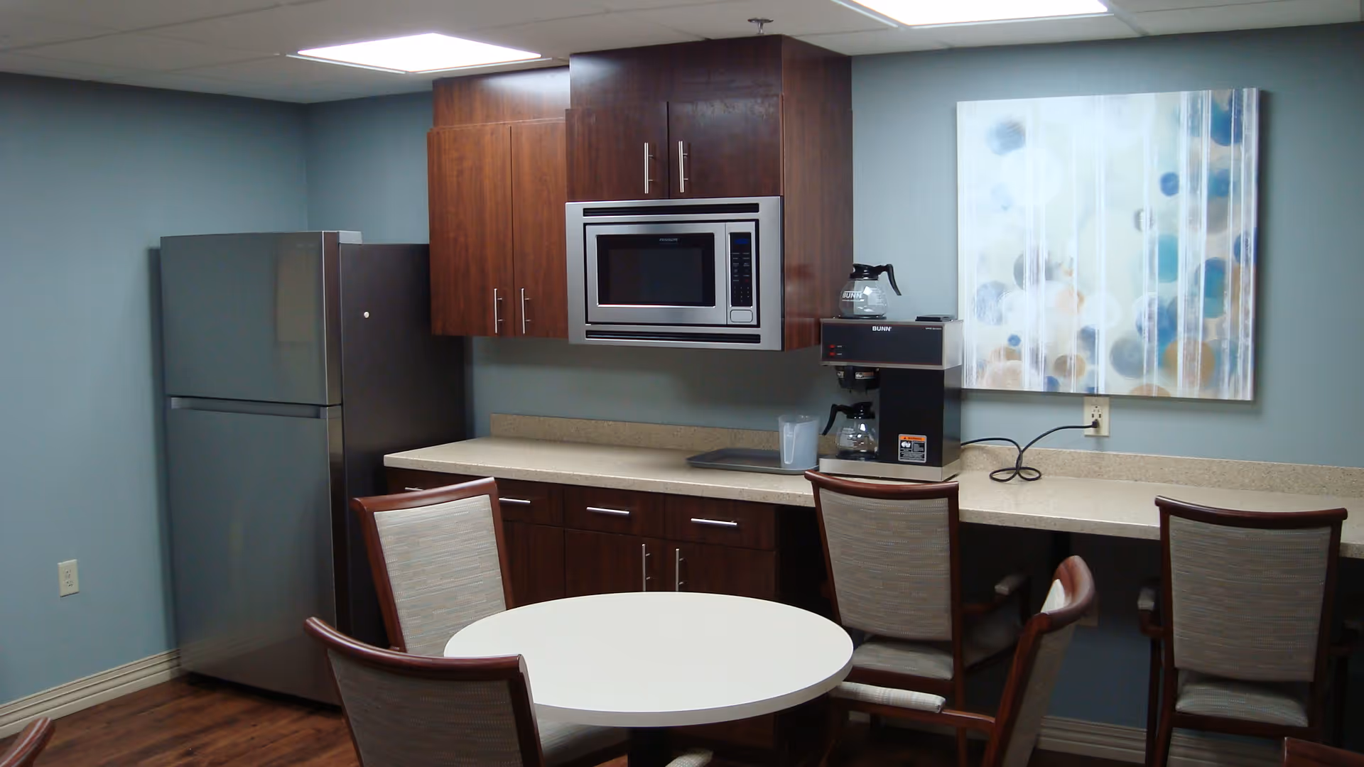 A small kitchen area with a round white table surrounded by four chairs. The kitchen has wooden cabinets, a stainless steel refrigerator, a microwave, and a coffee maker on the countertop. A colorful abstract painting is hung on the wall above the counter.