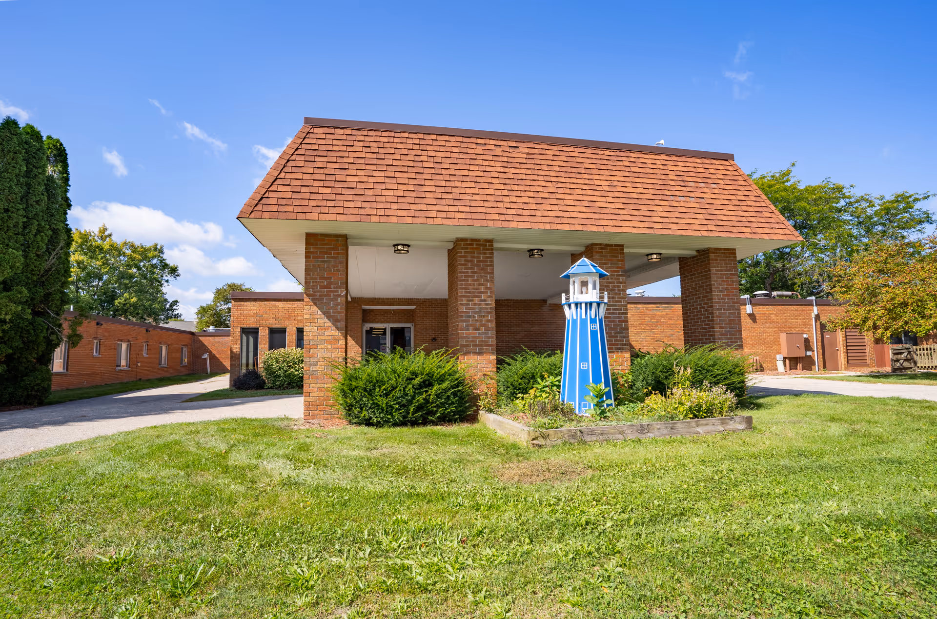 Exterior view of a brick building with a covered entrance supported by four brick pillars. In front of the entrance is a small garden bed with green shrubs and a decorative blue lighthouse structure. The building is surrounded by green grass and trees under a clear blue sky.