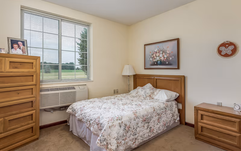 A cozy bedroom with a floral patterned bedspread on a bed against a cream-colored wall. There is a wooden dresser with framed photos on top, a wooden nightstand with a clock, a standing lamp, and a window with blinds showing a green outdoor view.
