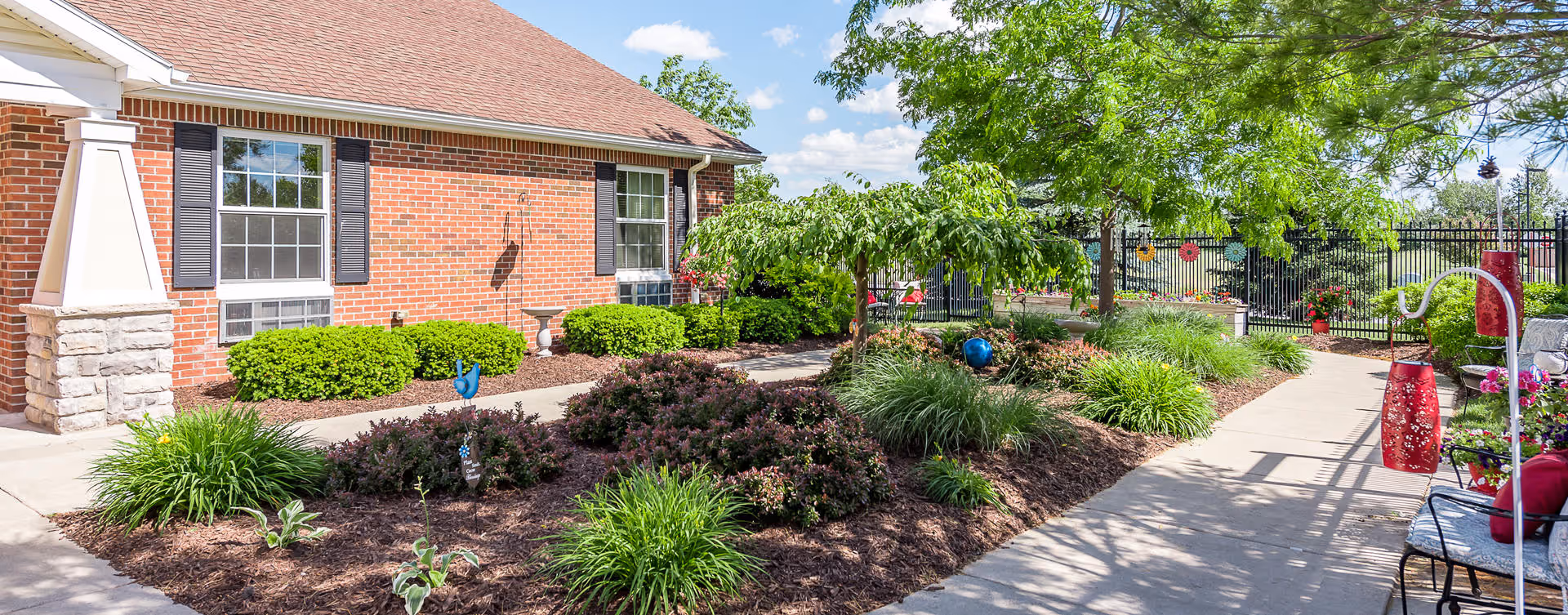 Outdoor garden area at Bickford of Midland featuring a brick building with windows, well-maintained landscaping with bushes, small trees, and decorative plants, a paved walkway, and outdoor seating with red cushions under a sunny blue sky.