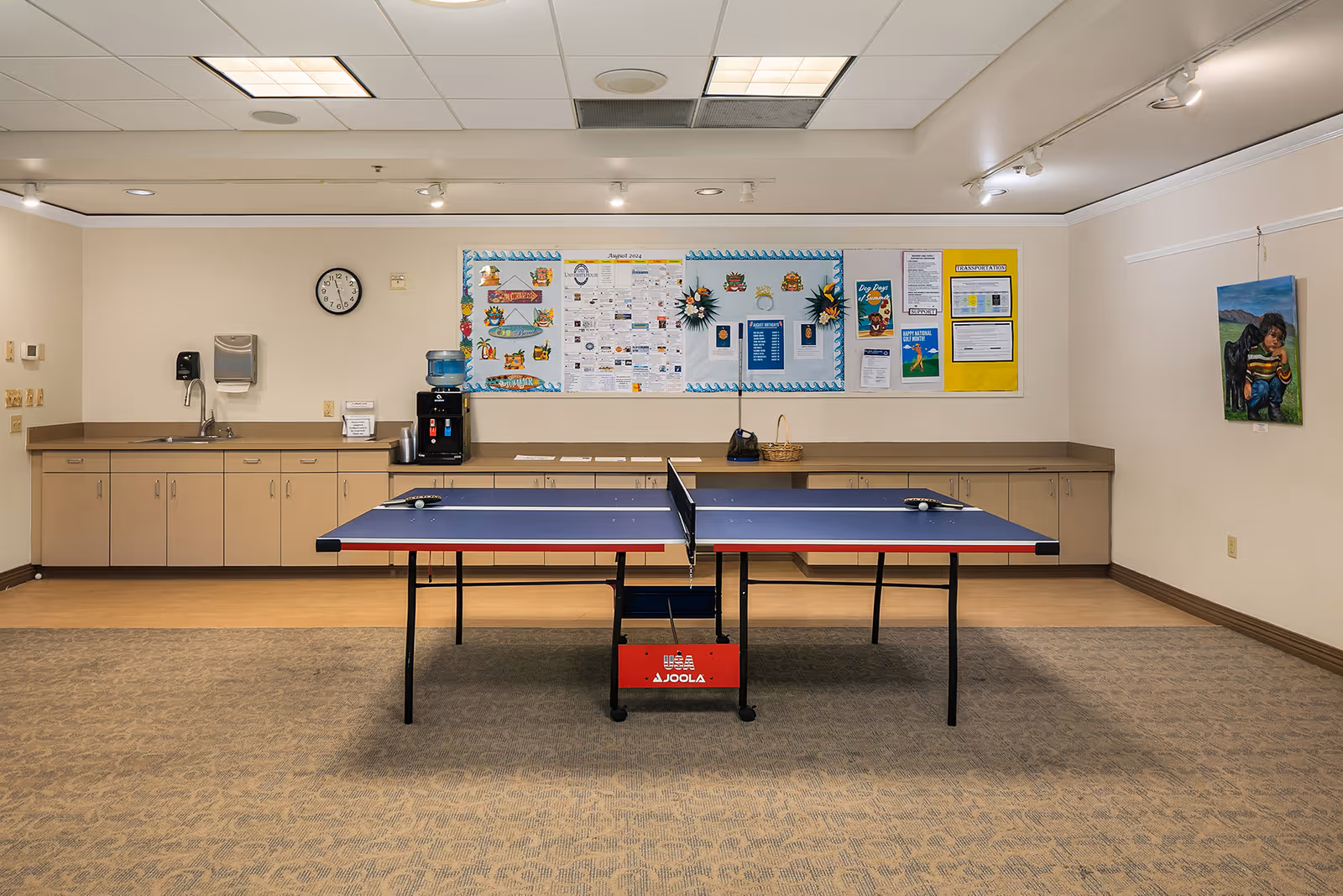 Indoor recreational room with a blue ping pong table in the center. The back wall has a long counter with cabinets underneath, a water dispenser, a sink, and a bulletin board with various notices and decorations. A clock is mounted on the wall above the counter. There is a painting of a child on the right wall. The room has a carpeted floor and ceiling lights.