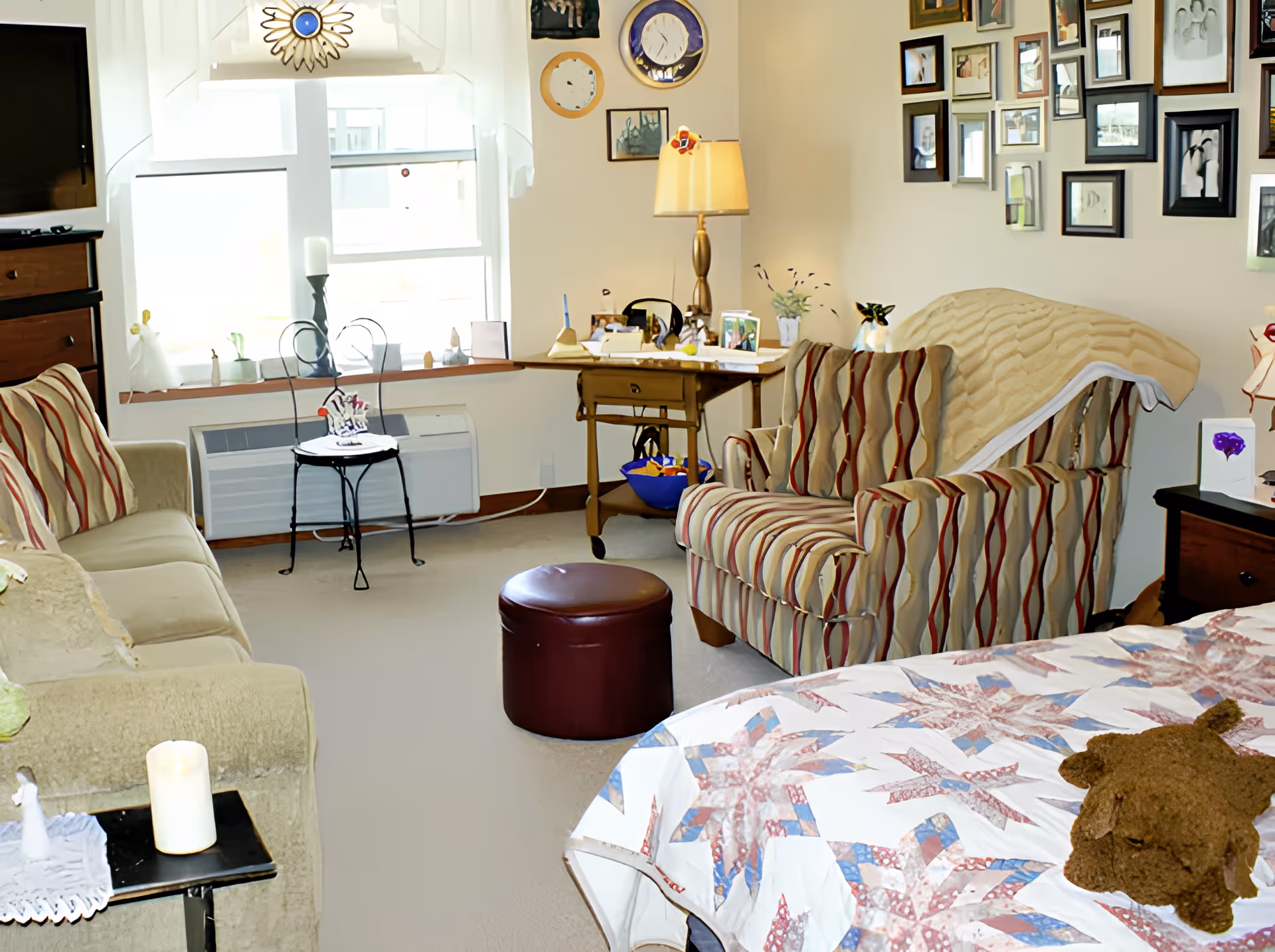 A cozy living space featuring a bed with a patterned quilt and a stuffed animal, a striped armchair with a blanket draped over it, a beige sofa with striped pillows, a small round ottoman, a wooden side table with a lamp, and a wall adorned with numerous framed pictures. A window with sheer curtains lets in natural light, and a small black metal chair is placed near the window.