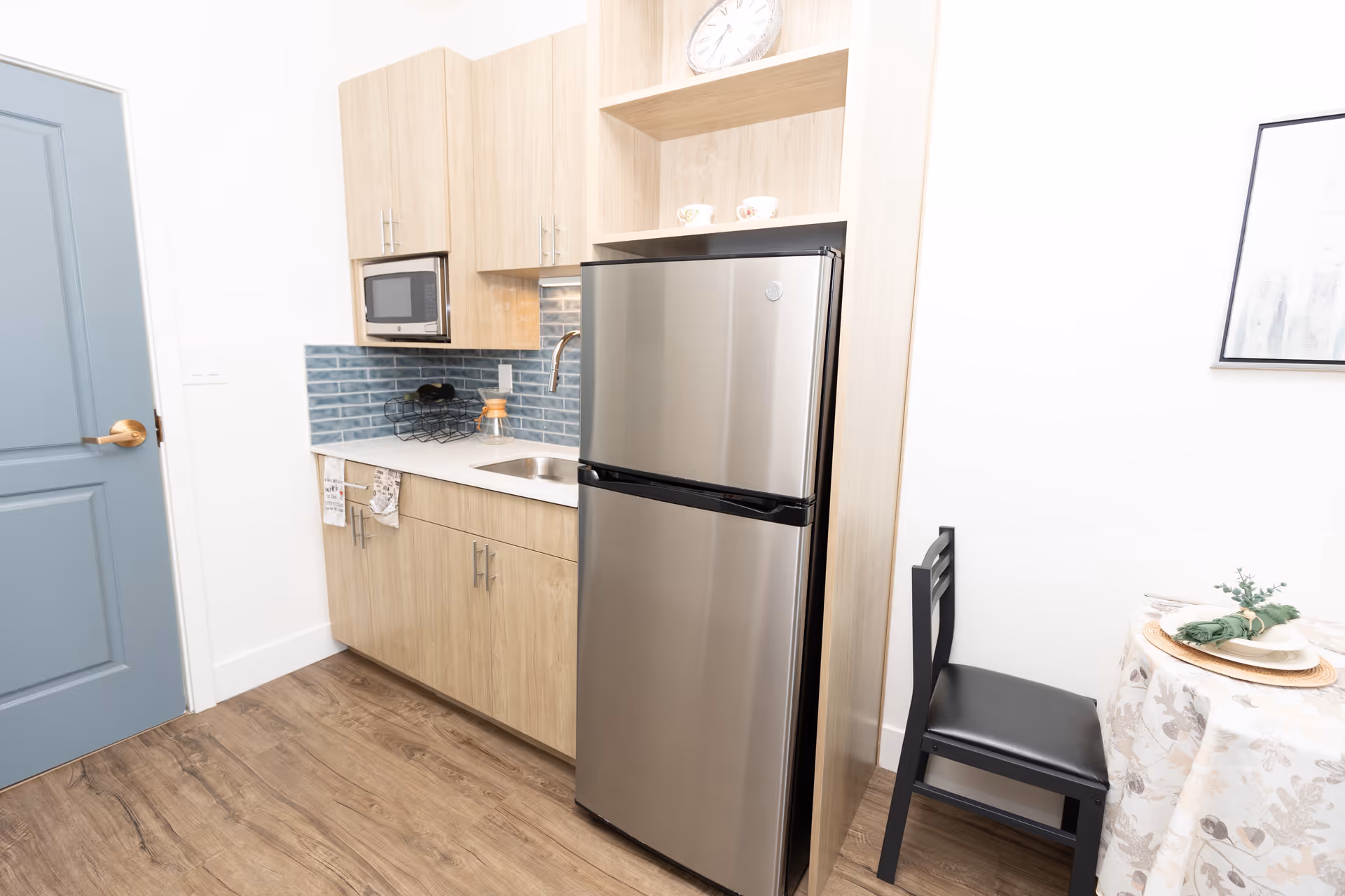 Small kitchenette with a stainless steel refrigerator, sink, microwave, blue tile backsplash, and a chair by a small table.
