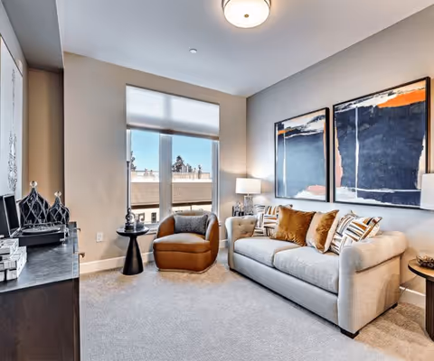 A cozy living room with a beige sofa adorned with multiple decorative pillows, a brown armchair, a small round black side table, and a lamp on a side table. Two large abstract paintings hang on the wall above the sofa. A window with a white shade lets in natural light, and a dark-colored cabinet with decorative items is visible on the left side.