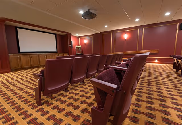 Interior view of a senior living facility's theater room with rows of maroon cushioned seats facing a large projection screen. The room has patterned carpet flooring, red walls with gold trim, and ceiling lights.