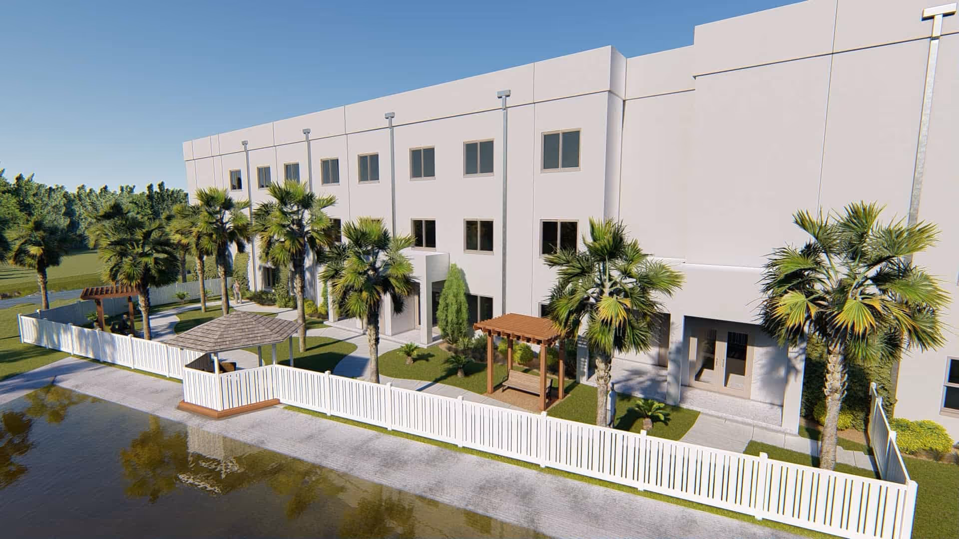 Exterior view of a multi-story building with several windows, surrounded by palm trees and a white fence. There are small wooden pergolas and a gazebo in a landscaped garden area next to a body of water under a clear blue sky.