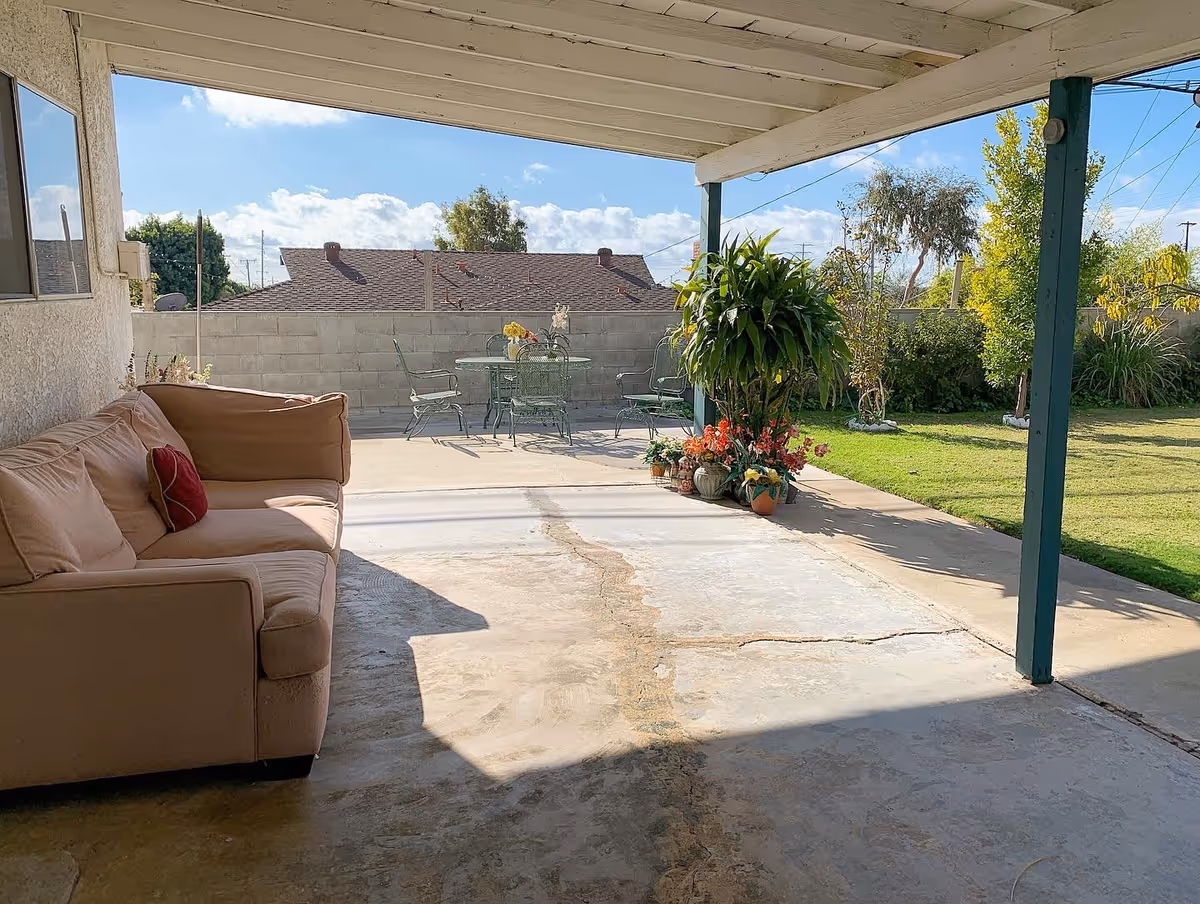 Covered backyard patio with a sofa, potted plants, and a metal outdoor dining set overlooking a lawn.