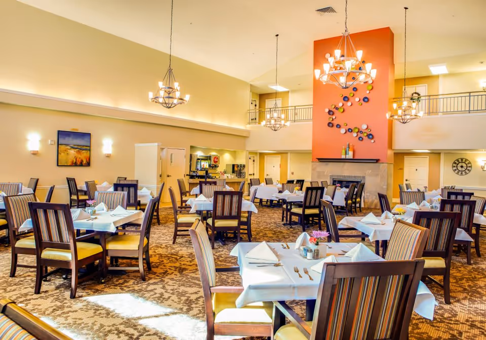 Spacious dining room with multiple set tables, chandeliers, and a colorful wall feature above a fireplace.