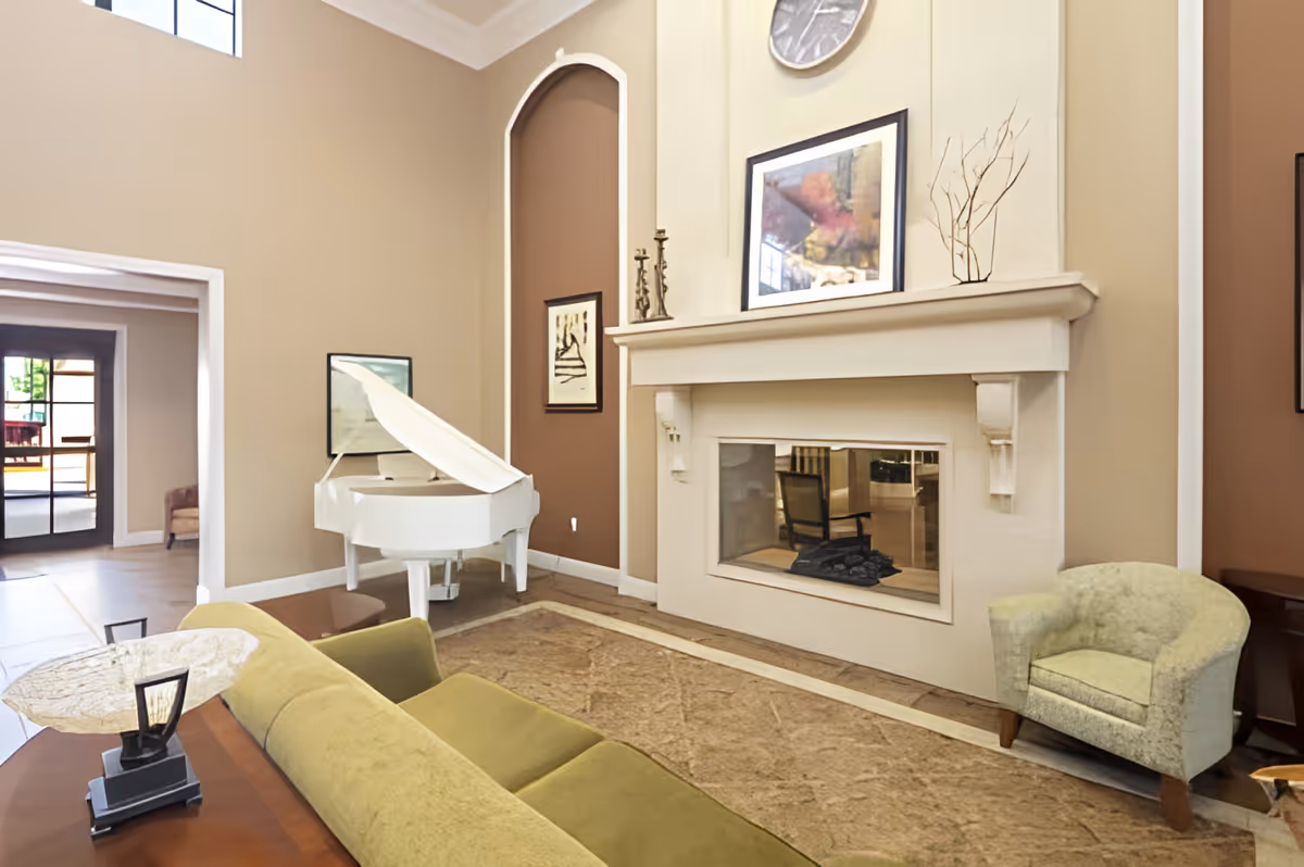 A cozy living room area with a green sofa, a light green armchair, a white grand piano, and a fireplace with a clock and framed artwork above it. The walls are painted in beige and brown tones, and there is a large window letting in natural light.