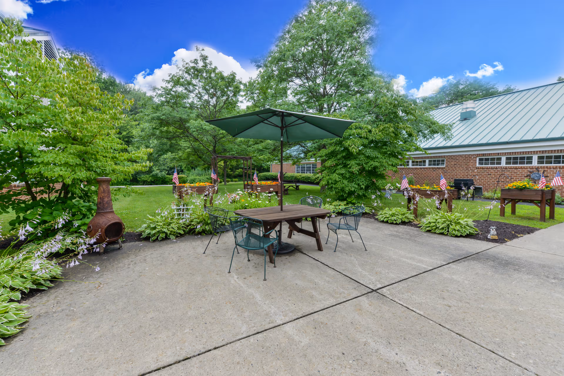 Outdoor patio area with a wooden table and four metal chairs under a large green umbrella. Surrounding the patio are green trees, plants, and flower beds with small American flags. A brick building with a green metal roof is visible in the background under a bright blue sky with some clouds.
