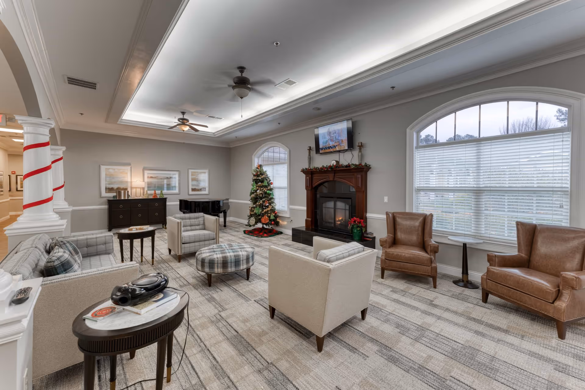 A spacious and well-lit living room with a Christmas tree decorated for the holidays, a fireplace with a TV mounted above it, several armchairs and a sofa arranged around a plaid ottoman, a grand piano in the corner, and large windows with blinds letting in natural light.