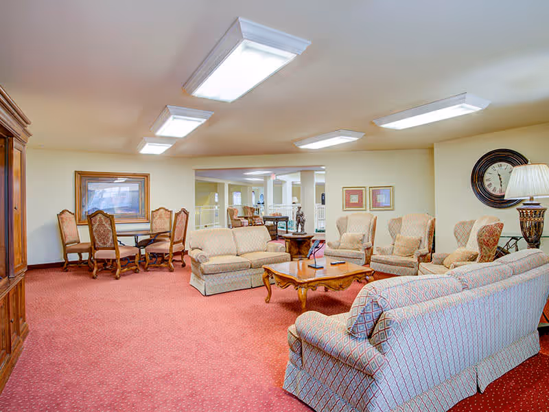 A spacious living room area in an assisted living facility with patterned sofas and armchairs arranged around a wooden coffee table. The room has a red carpet, cream-colored walls, and several framed artworks and a large clock on the walls. There is also a wooden dining table with chairs in the background and bright ceiling lights.