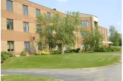 Exterior view of a three-story brick building with multiple windows, surrounded by green grass, trees, and a paved walkway under a clear sky.