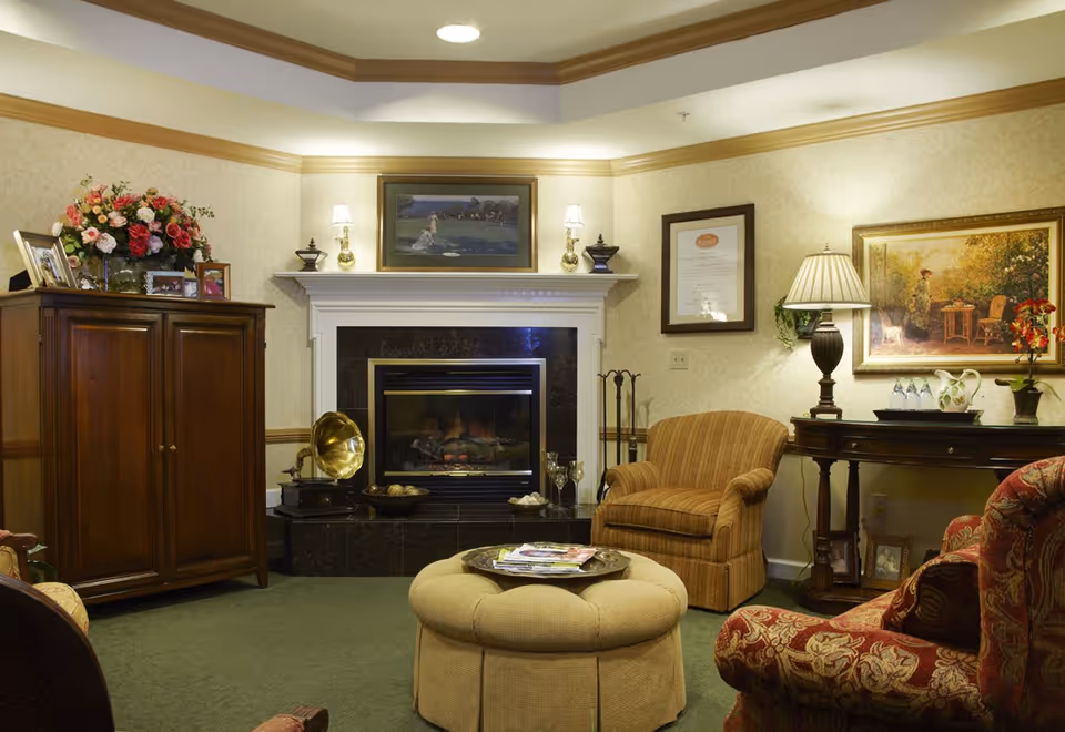 A cozy living room with a fireplace in the center, flanked by two wall sconces and decorative items on the mantel. There is a wooden cabinet with framed photos and a flower arrangement on the left. In front of the fireplace is a round beige ottoman with magazines on a tray. To the right, there is a striped armchair and a wooden console table with a lamp, decorative items, and framed pictures underneath. The walls are decorated with framed artwork and the room has a warm, inviting ambiance.