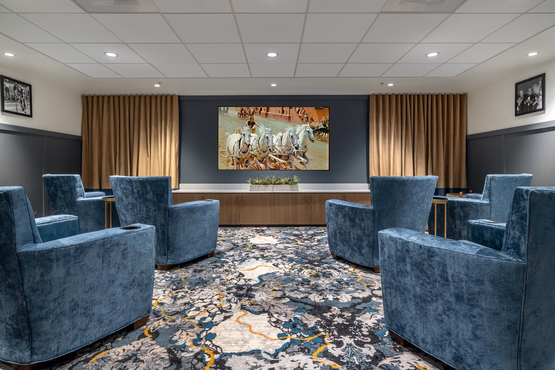 A cozy senior living common area with six blue upholstered armchairs arranged in two rows facing a large flat-screen TV mounted on a dark accent wall. The TV displays an image of a horse-drawn carriage race. The room has patterned carpet with blue, gray, and yellow tones, beige curtains on either side of the TV, and framed black and white photographs on the walls.