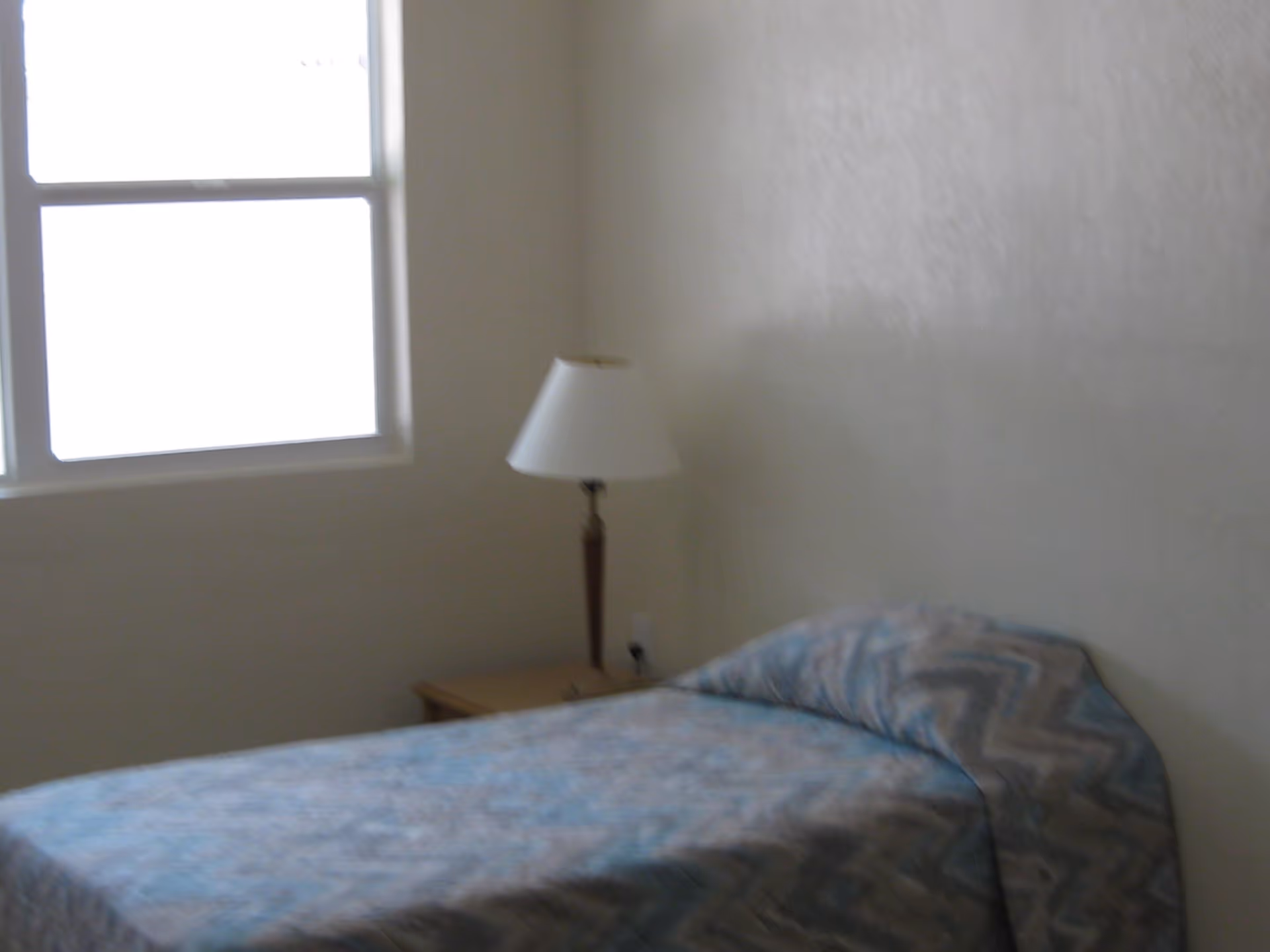 A simple bedroom with a single bed covered in a blue and gray patterned bedspread. Next to the bed is a wooden nightstand with a white lampshade on a lamp. There is a window on the left wall letting in natural light.